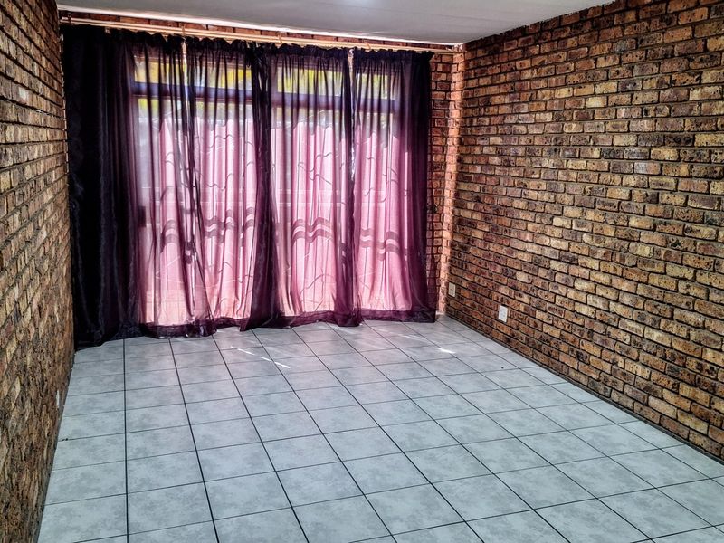 An empty living room featuring exposed brick walls and ceramic floor tiles, with a large sliding glass door providing natural light.