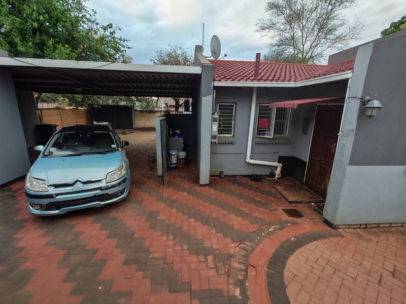 Front exterior view of the property featuring a paved driveway, attached carport, and tiled roof structure with visible maintenance required on exterior finishes.