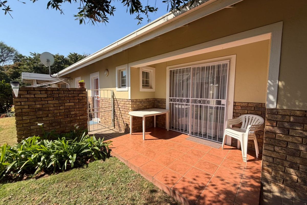 A covered patio area featuring tiled flooring, brick accents, and a sliding glass door with security gate, overlooking a small garden space.