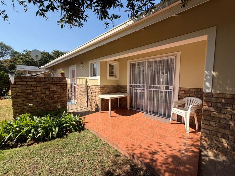 A covered patio area featuring tiled flooring, brick accents, and a sliding glass door with security gate, overlooking a small garden space.