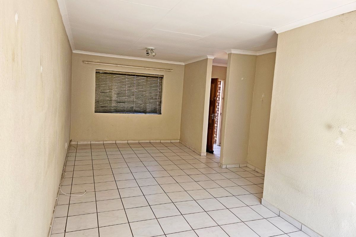 An empty, open-plan living area featuring neutral tiled flooring, coved ceilings, and a window fitted with venetian blinds.