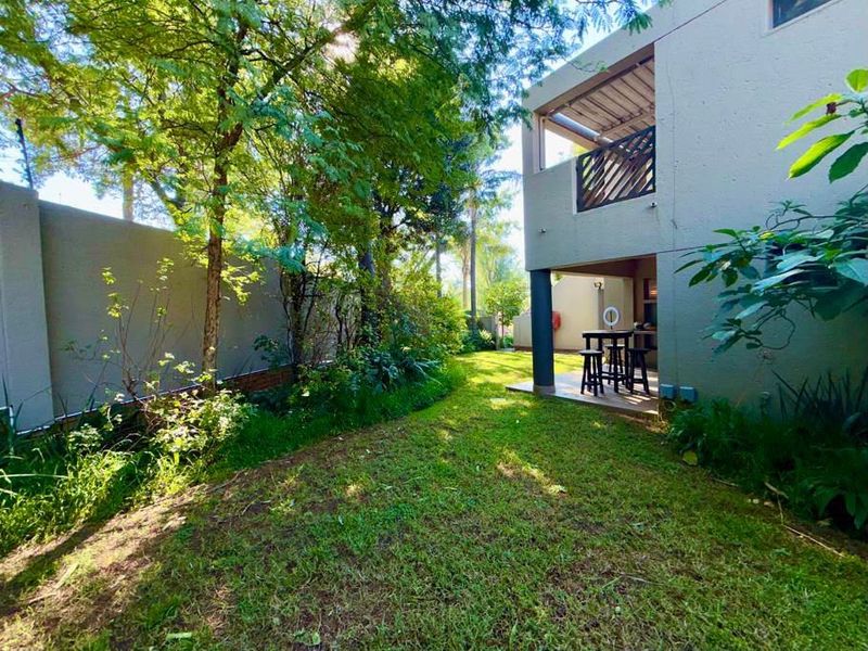 A private rear garden featuring a lush lawn, mature trees, a covered patio area, and an upper-level balcony attached to the main dwelling.