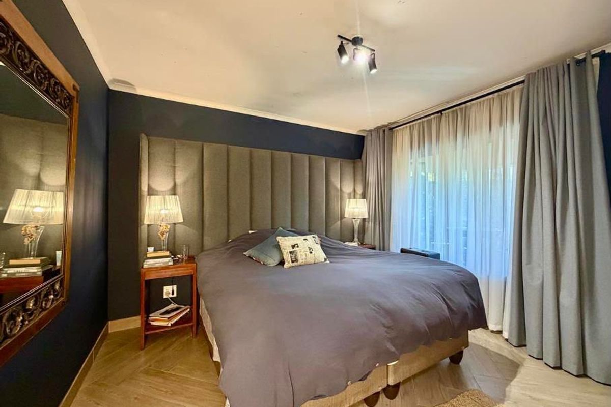 A well-appointed bedroom featuring dark accent walls, a large upholstered headboard, herringbone flooring, and floor-to-ceiling curtains providing access to the exterior.