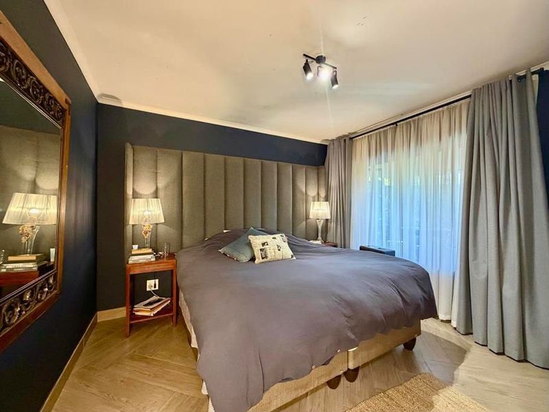 A well-appointed bedroom featuring dark accent walls, a large upholstered headboard, herringbone flooring, and floor-to-ceiling curtains providing access to the exterior.