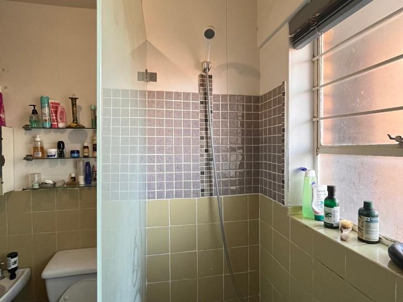 A functional bathroom featuring a bathtub with a shower mixer, glass screen, toilet, and tiled wall finishes with built-in shelving.