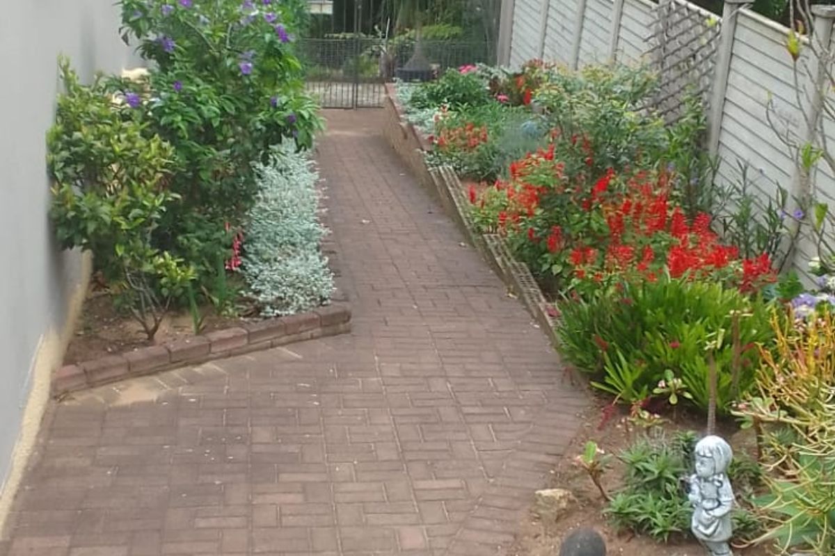 A well-maintained side garden path featuring brick paving, established garden beds, and secure perimeter fencing.