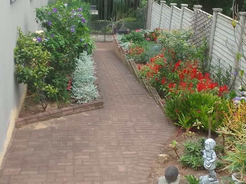 A well-maintained side garden path featuring brick paving, established garden beds, and secure perimeter fencing.