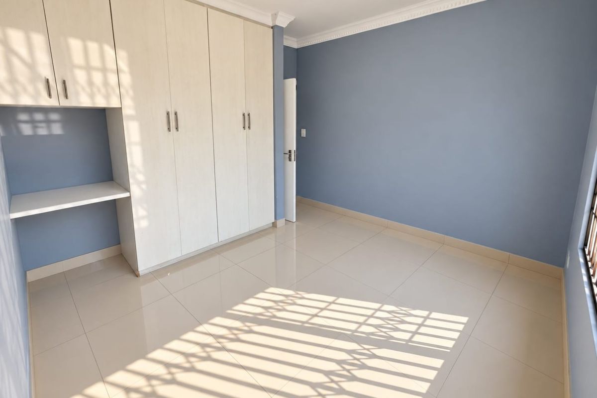 A bright, empty bedroom featuring neutral tiled flooring, blue walls, and ample built-in storage with an integrated desk area.