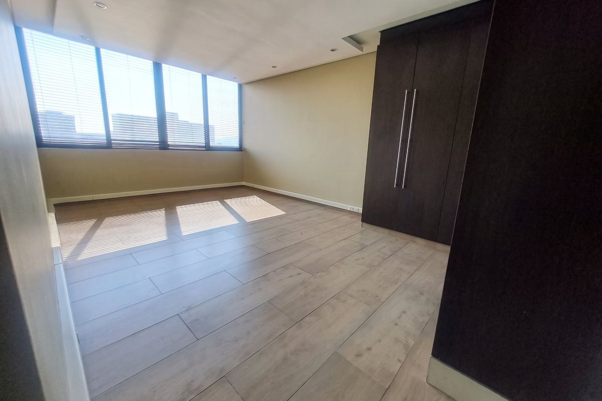 A spacious, empty bedroom featuring modern laminate flooring, built-in dark wood cupboards, recessed ceiling lighting, and large windows with fitted venetian blinds.