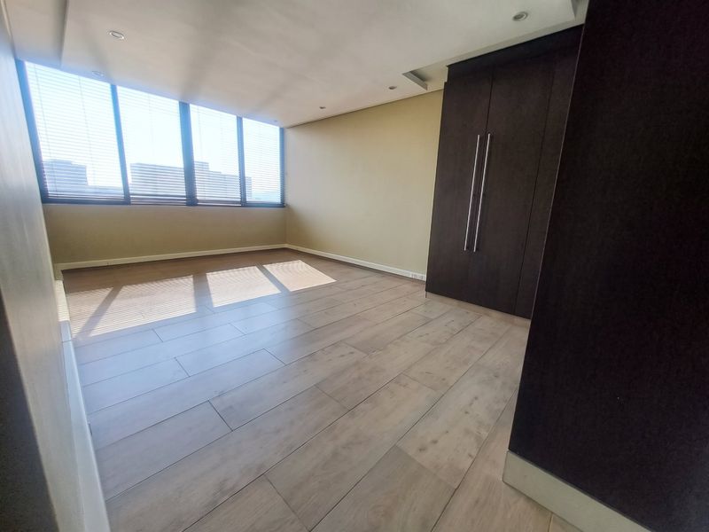 A spacious, empty bedroom featuring modern laminate flooring, built-in dark wood cupboards, recessed ceiling lighting, and large windows with fitted venetian blinds.