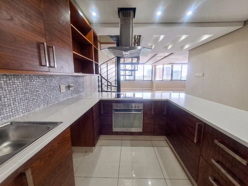 Modern kitchen featuring dark wood cabinetry, stone countertops, mosaic backsplash, and integrated stainless steel appliances, adjacent to an open-plan living area with spiral staircase.