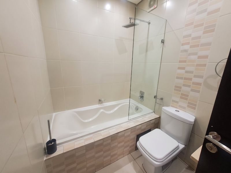A clean, modern bathroom featuring a built-in bathtub with a glass shower screen, rainfall shower head, and a standard toilet with neutral tiled finishes.
