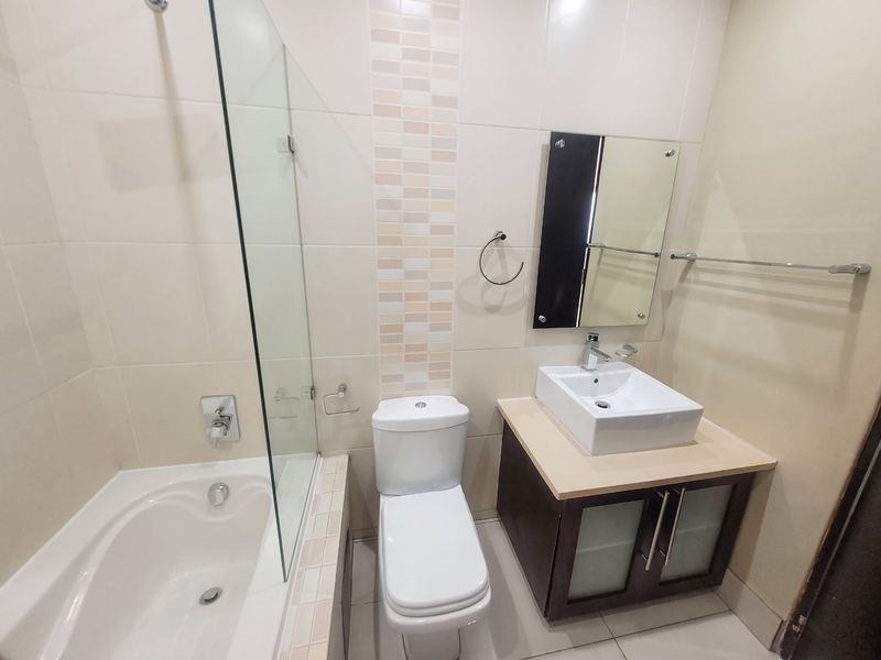 A clean, modern bathroom featuring a built-in vanity with a vessel sink, a toilet, and a bathtub with a glass shower screen.