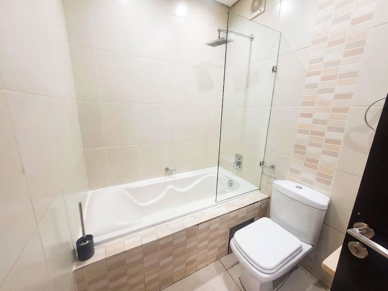 A clean, modern bathroom featuring a built-in bathtub with a glass shower screen, rainfall shower head, and a standard toilet with neutral wall tiling.