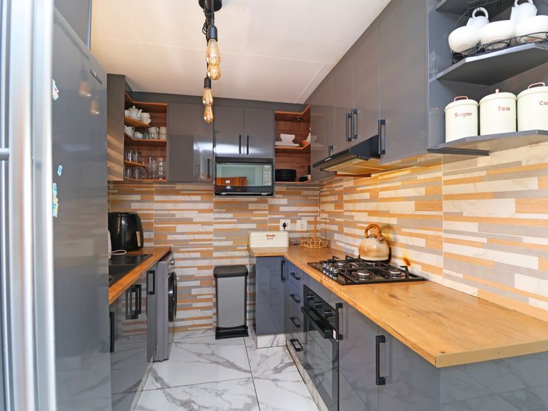 Modern galley kitchen featuring grey high-gloss cabinetry, wooden countertops, gas hob, built-in oven, and stylish stone-look tile backsplash.