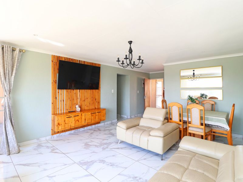 Open-plan living and dining area featuring marble-look floor tiles, a custom wooden TV feature wall, and a classic chandelier.