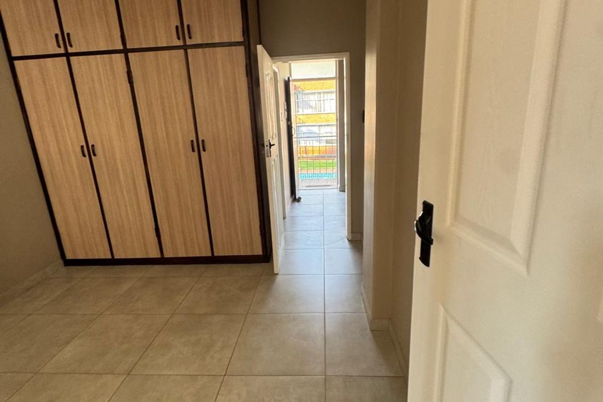 A clean, empty bedroom featuring neutral tiled flooring and built-in wooden cupboards, with a view through the doorway toward an exterior balcony area.