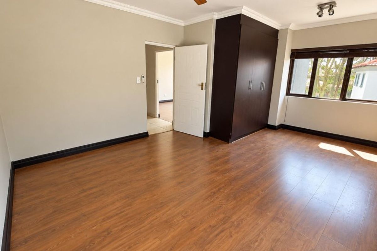 A spacious, empty bedroom featuring laminate flooring, a built-in dark wood wardrobe, and large windows with blinds.