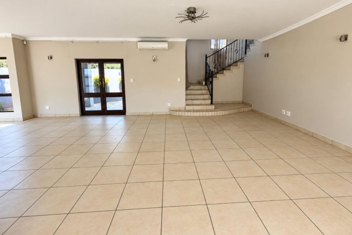 Spacious, open-plan living area featuring neutral tiled flooring, a staircase with wrought iron balustrades, and a wall-mounted air conditioning unit.