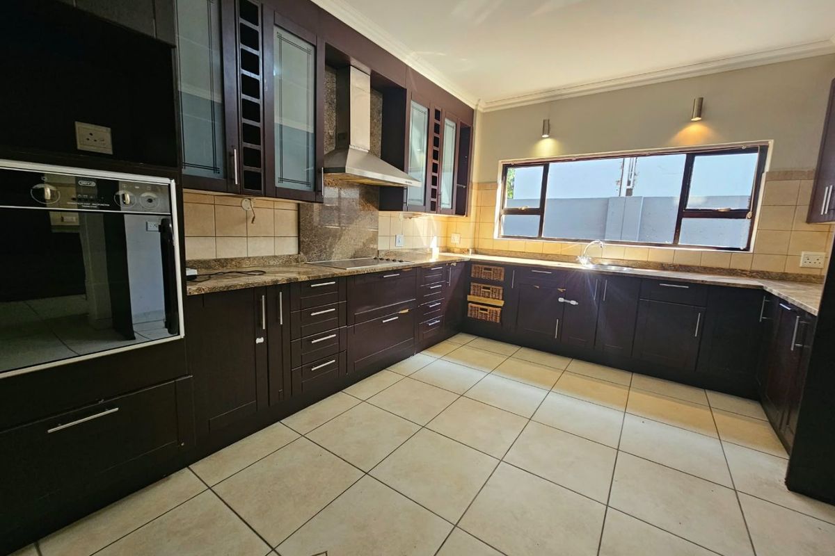 A spacious kitchen featuring dark wood cabinetry, granite countertops, built-in oven, stainless steel extractor fan, and tiled flooring with ample natural light.