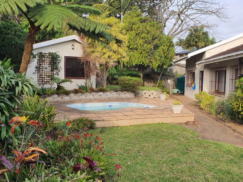 A private backyard area featuring a swimming pool with a paved deck, surrounded by established garden landscaping and a secondary outbuilding structure.