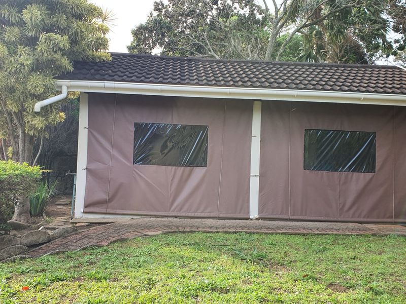 A covered outdoor patio area featuring a tiled roof, paved flooring, and protective canvas blinds, set within a landscaped garden environment.