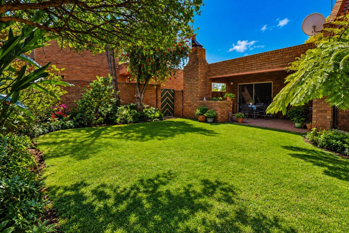 A well-maintained rear garden featuring a lush lawn, established trees, and a paved patio area with a built-in brick fireplace.