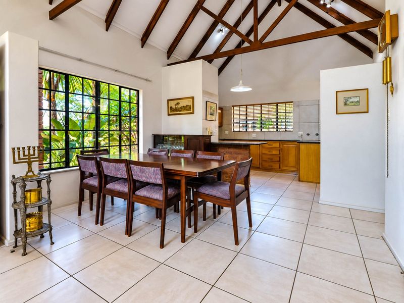 Spacious open-plan dining and kitchen area featuring high vaulted ceilings with exposed timber trusses, tiled flooring, and large windows overlooking the garden.
