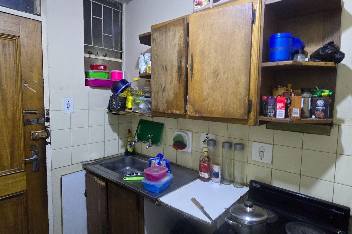 A compact kitchen featuring dated wooden cabinetry, a stainless steel sink, tiled splashback, and an electric stove, requiring cosmetic updates and refurbishment.