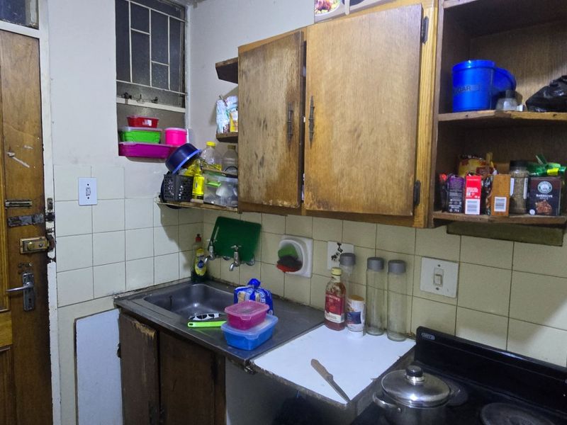 A compact kitchen featuring dated wooden cabinetry, a stainless steel sink, tiled splashback, and an electric stove, requiring cosmetic updates and refurbishment.