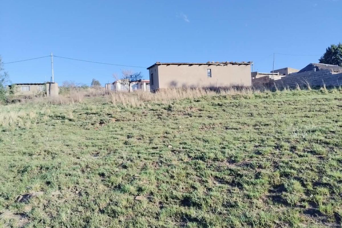 A view of a sloped plot of land featuring an unfinished structure with visible roof damage and surrounding boundary walls.