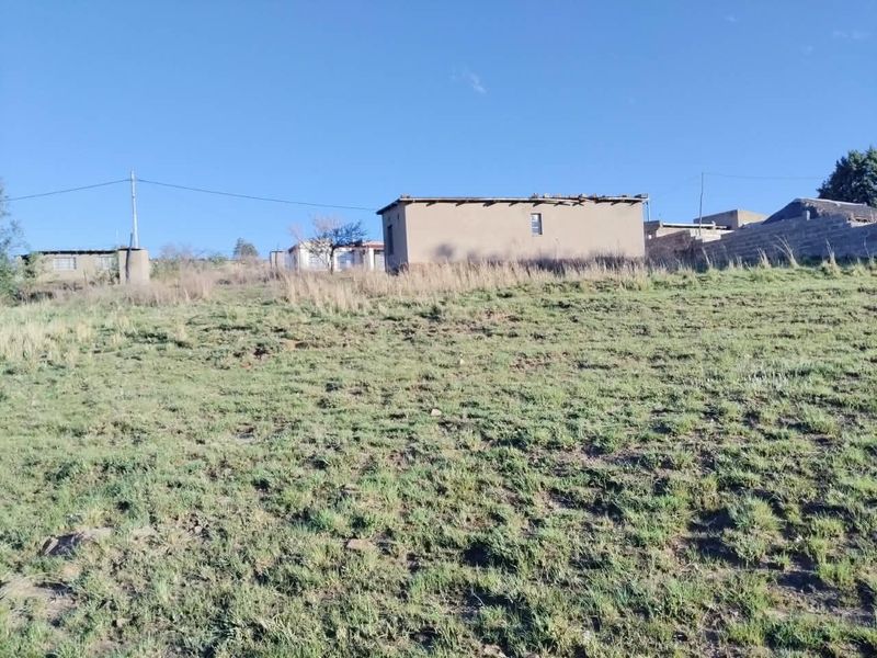 A view of a sloped plot of land featuring an unfinished structure with visible roof damage and surrounding boundary walls.