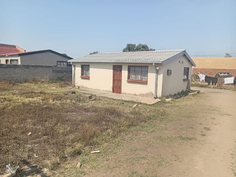 A detached single-story house featuring a corrugated metal roof, security gates, and burglar bars, situated on a large, unmaintained plot.