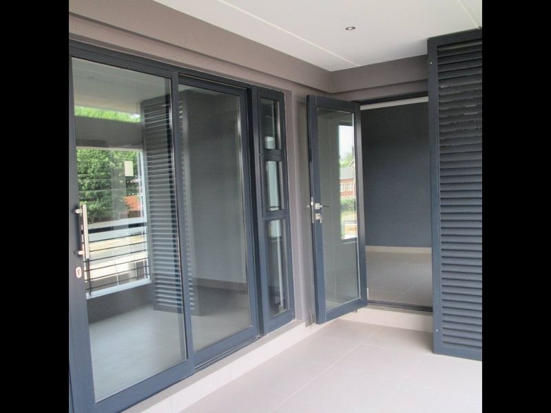 A modern, covered balcony featuring tiled flooring, recessed lighting, and dark-framed glass doors with integrated louvred shutters.
