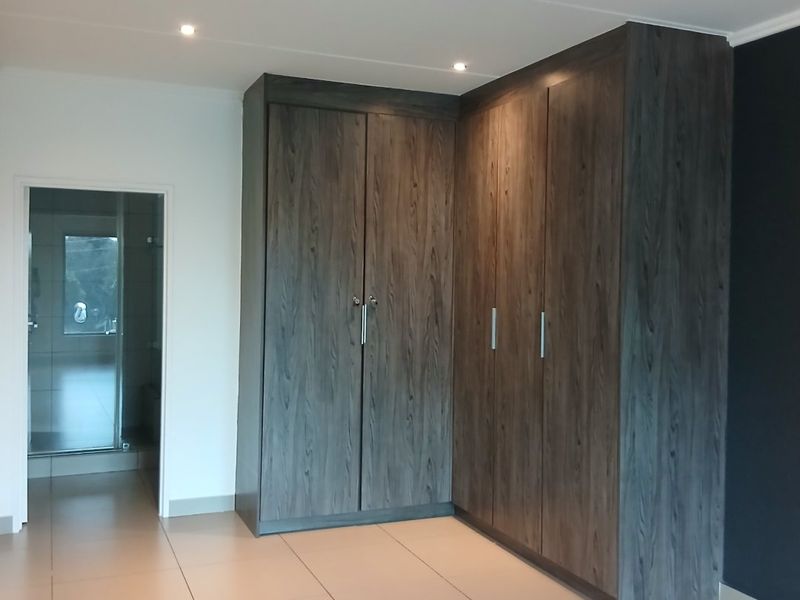 A spacious bedroom featuring modern dark wood built-in cupboards, neutral tiled flooring, and recessed downlighting, with an adjacent en-suite bathroom visible.