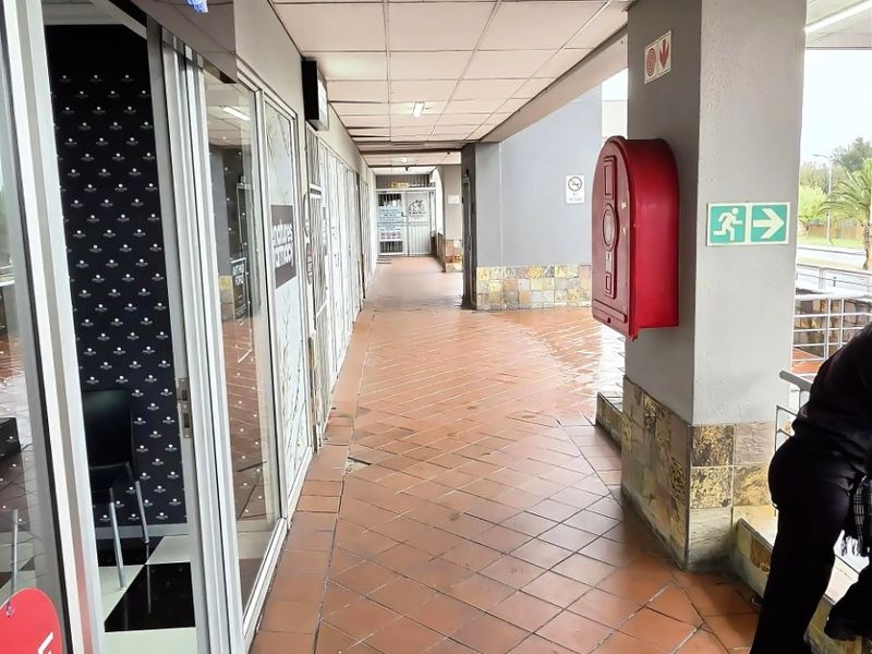 Covered commercial walkway featuring tiled flooring, stone-clad pillars, and glass storefront access points, equipped with a fire hose reel and emergency signage.