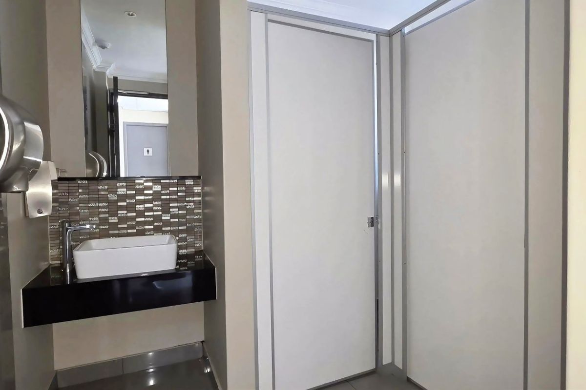 A clean, modern commercial bathroom featuring a vessel sink, wall-mounted faucet, tiled backsplash, and partitioned toilet cubicles.