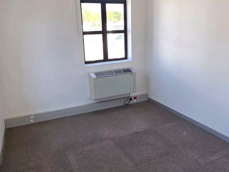 A clean, empty office space featuring carpeted flooring, a suspended ceiling with recessed lighting, and a wall-mounted air conditioning unit.