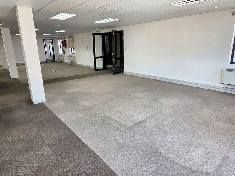 Spacious open-plan commercial office space featuring neutral carpeted flooring, suspended ceilings with recessed lighting, and integrated air conditioning units.