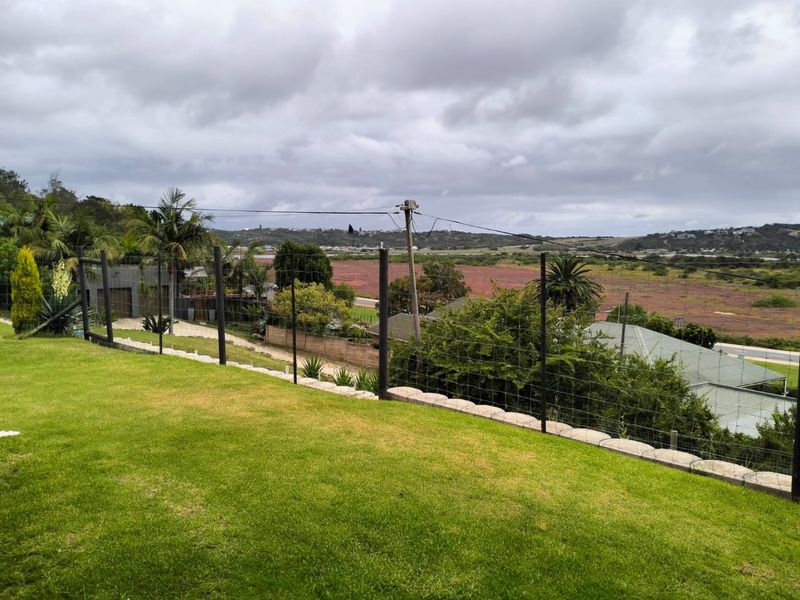 An elevated view overlooking a grassy lawn, wire mesh fencing, and a retaining wall, with a distant landscape and overcast sky.