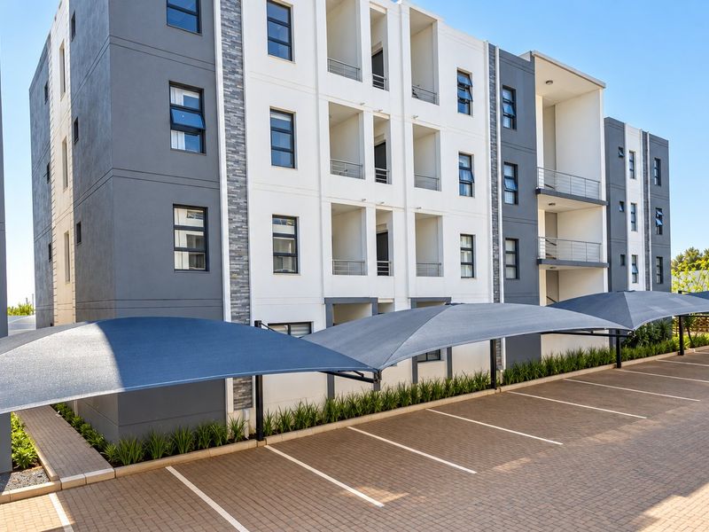 Modern multi-story apartment building exterior featuring a paved parking area with shade cloth carports and well-maintained landscaped garden beds.
