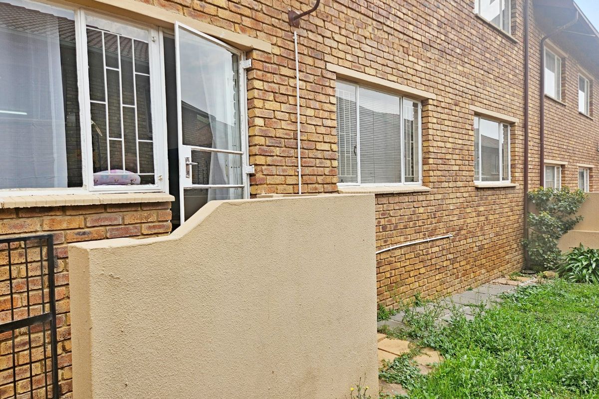 Rear exterior view of a face brick townhouse featuring security bars on windows, a small paved area, and a concrete privacy wall.
