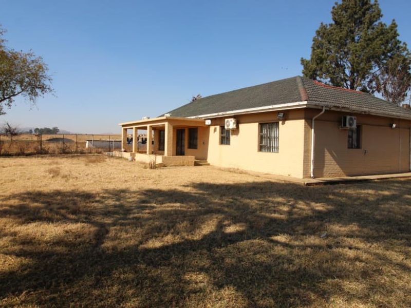 Rear view of a single-story brick farmhouse featuring a covered patio, tiled roof, and installed air conditioning units set on a large rural plot.