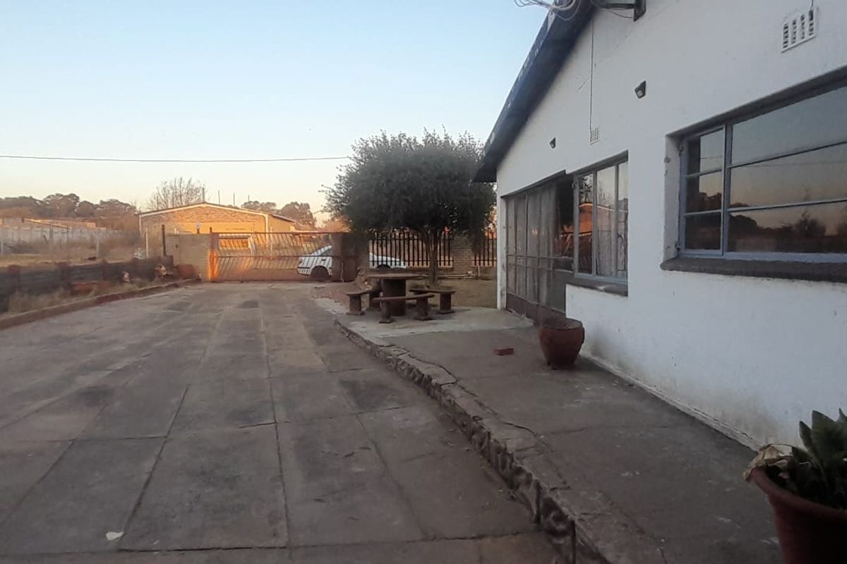 Rear exterior view of the property featuring a large paved driveway, concrete patio area, and perimeter security fencing.