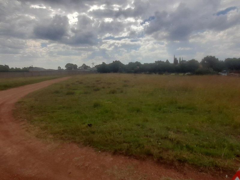 A wide-angle view of a vacant residential plot featuring a dirt driveway and perimeter boundary wall under a cloudy sky.