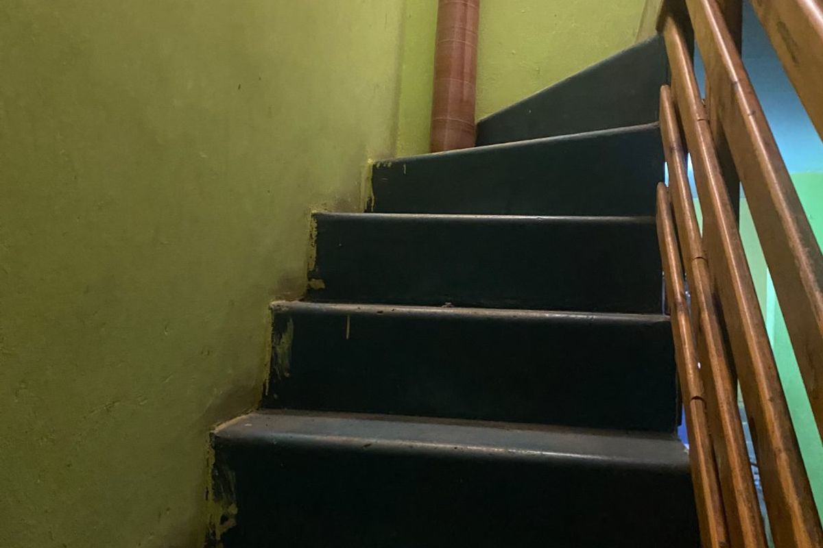 A narrow interior staircase featuring dark painted wooden steps and a simple wooden handrail, showing signs of wear and dated wall finishes.