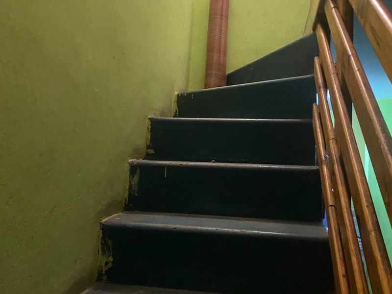 A narrow interior staircase featuring dark painted wooden steps and a simple wooden handrail, showing signs of wear and dated wall finishes.