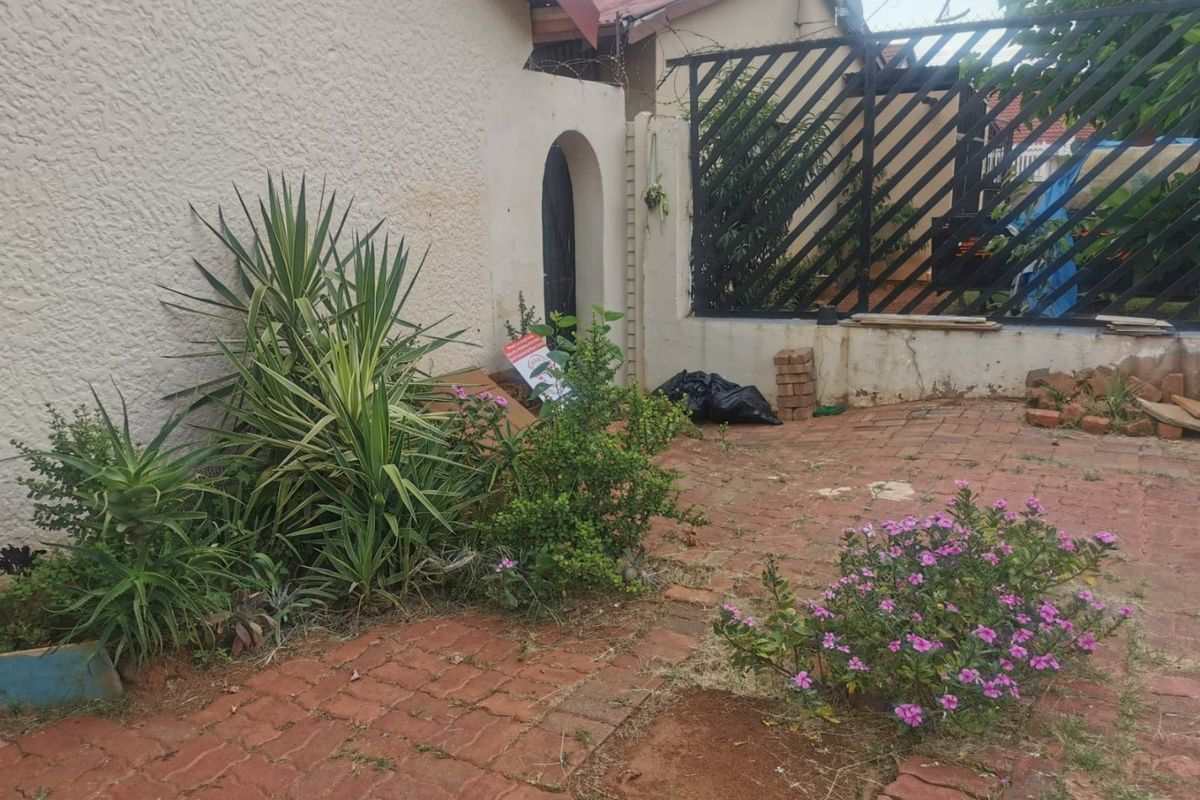 A rear exterior area featuring brick paving, a metal security gate, and a plastered boundary wall with an arched entrance.