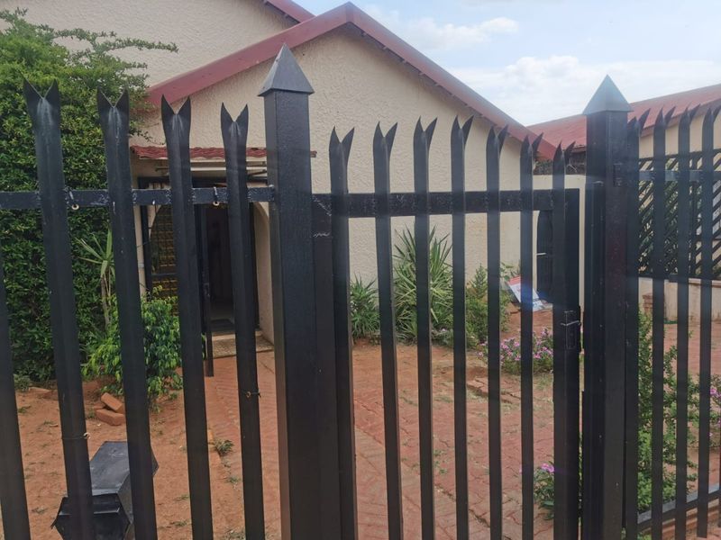 Front view of the property featuring a secure metal fence and gate, paved entrance path, and a stucco facade with a tiled roof.