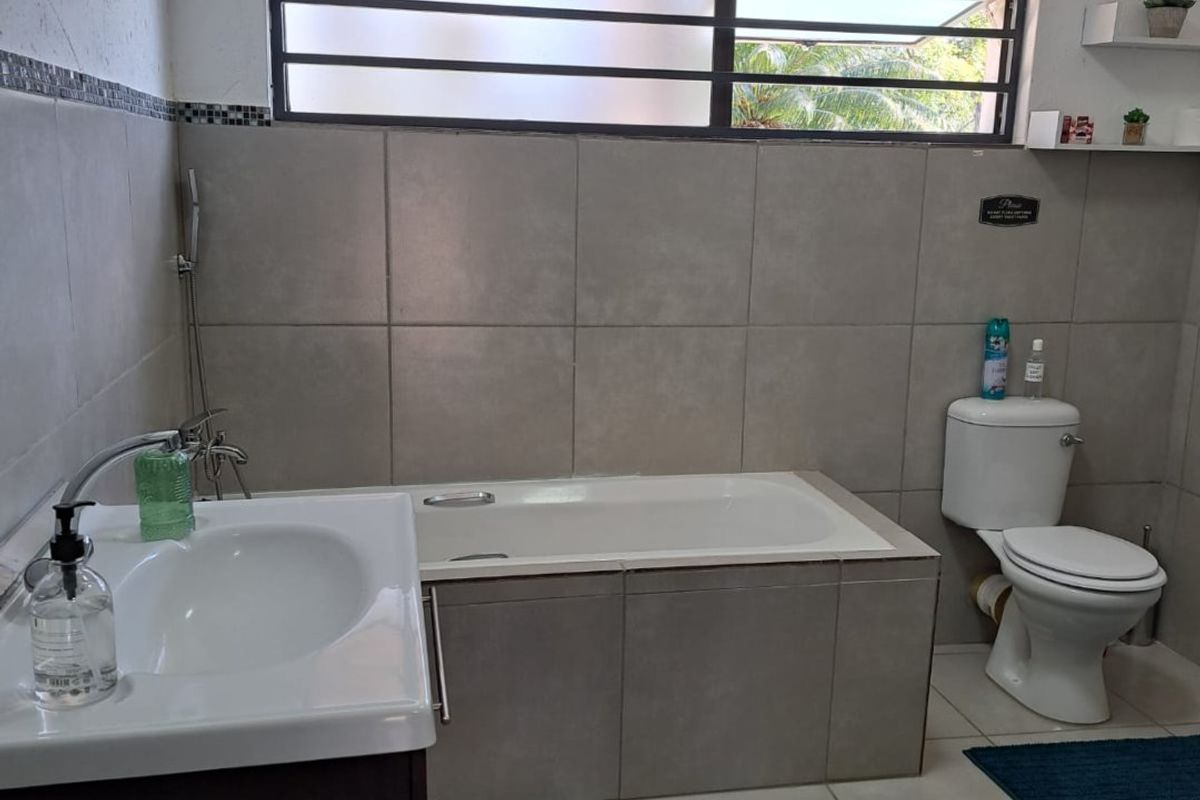A functional bathroom featuring a built-in bathtub, vanity basin, and toilet, finished with neutral wall and floor tiling and a horizontal window.